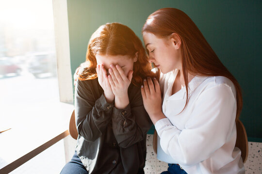 Young woman crying in a cafe covers her face with her hands. Female model hugs her friend.