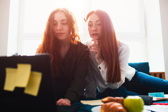 Two red-haired students study at home or prepare for exams. Young women doing homework in a dormitory bed near the window. There are notebooks, food books, a tablet and laptop and documents