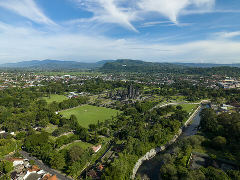 Aerial view of Prambanan Temple complex surrounded by lush green fields, a winding river, and urban landscape under a blue sky with wispy clouds in Prambanan, Daerah Istimewa Yogyakarta, Indonesia.