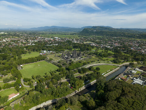 Aerial view of Prambanan Temple featuring ancient stone spires, lush green parkland, a winding river, and distant mountains under a blue sky in Prambanan, Daerah Istimewa Yogya, Indonesia.