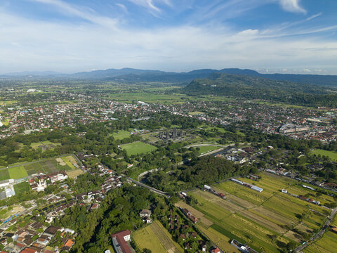 Aerial view of Prambanan Temple complex surrounded by lush green trees, agricultural fields, and urban settlements under a blue sky with mountains in Prambanan, Daerah Istimewa Yogya, Indonesia.