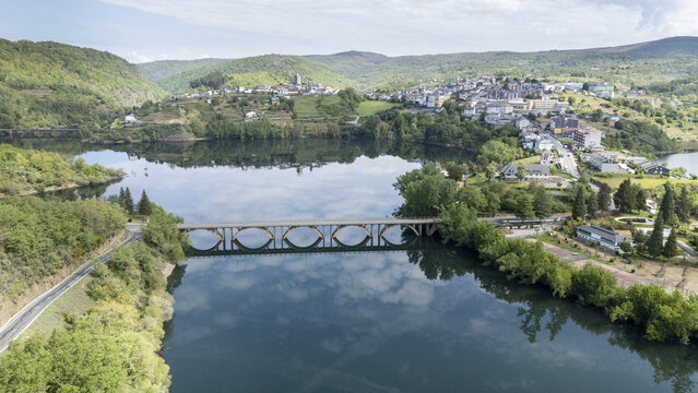 Aerial view of a stone arched bridge crossing a calm river with reflections of the sky and the town of Viana do Bolo on a green hill Viana do Bolo, Province of Ourense, Spain.