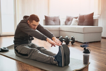 Young ordinary man go in for sport at home. Beginner or amateur in workout activity sit on mat and stretch to his toes. Hardworking guy exercising in apatment alone. © Vlad