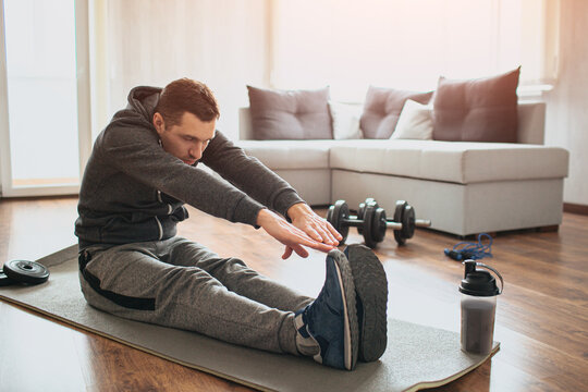 Young ordinary man go in for sport at home. Beginner or amateur in workout activity sit on mat and stretch to his toes. Hardworking guy exercising in apatment alone.