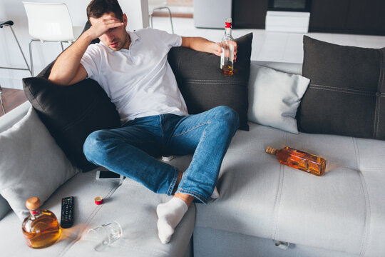Young man watch tv in his own apartment. Alcoholic sit on sofa amoing opened bottles with whiskey. Suffer from hangover or headache. Alone with problems. Desparate and hopeless.