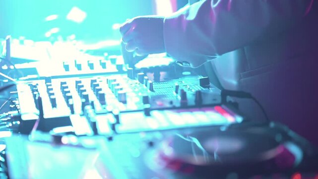 DJ operating mixing console with knobs faders and illuminated pads under neon lighting with shallow depth of field and dynamic reflections, slight camera roll movement, close up shot in slow motion