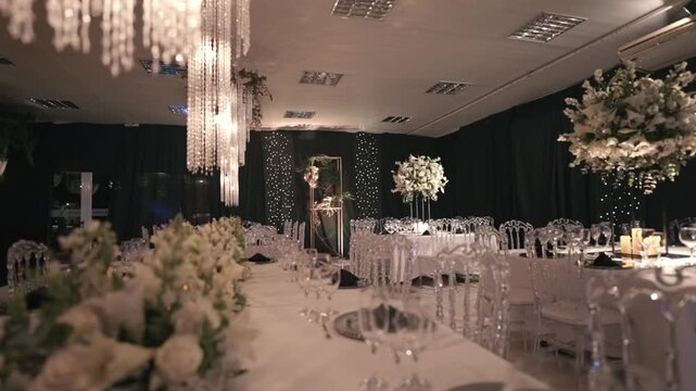 Luxurious wedding reception setup featuring a long dining table, clear ghost chairs, elegant white floral centerpieces, and sparkling crystal chandeliers.