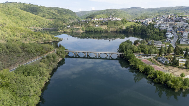 Aerial view of an arched bridge crossing a calm reservoir surrounded by lush green hills and the historic town of Viana do Bolo, Province of Ourense, Spain.