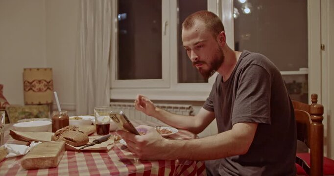 A casual scene of a young man intently scrolling on his smartphone at the dining table. He is having a quiet meal at home, immersed in his digital world.