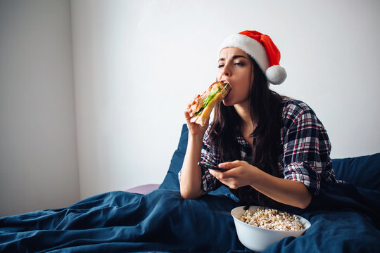 Young beautiful woman in morning bed at home. Christmas time or ney year period. Female model in red hat watching tv or movie. Eat big sandwich and popcorn. Use remote control.