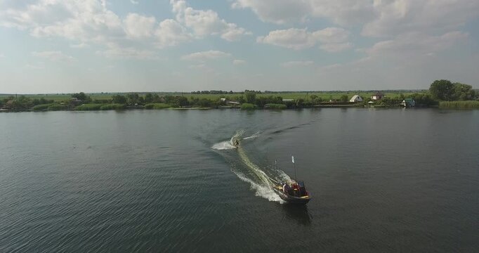 Mykolaiv Region, Ukraine - June 20, 2020: Water skier towed by motorboat on wide calm river. Foam wake curves across dark surface. Green reeds, riverside dachas and flat fields on far bank. Summer sky