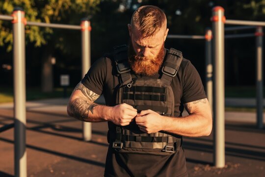 Bearded man puts on weighted training vest in park. Athletic person prepares for outdoor strength workout in early morning sunlight. Tattoos visible on muscular arm.