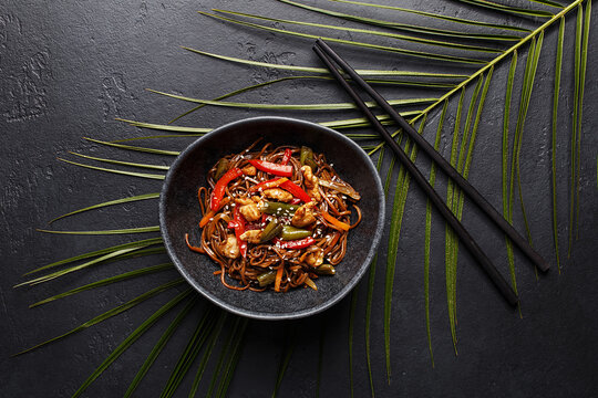 Stir-fry soba asian noodles with chicken, vegetables and sesame seeds in black ceramic bowl on black stone background with tropical palm leaf and chopsticks. Overhead view