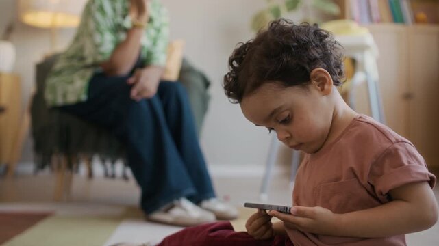 Handheld shot of adorable Black toddler using smartphone and sitting on floor while mother talking on mobile phone in background