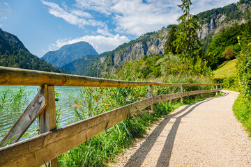 Fototapeta premium Walking path by the lake surrounded by mountains and trees on a sunny day with clouds in the sky