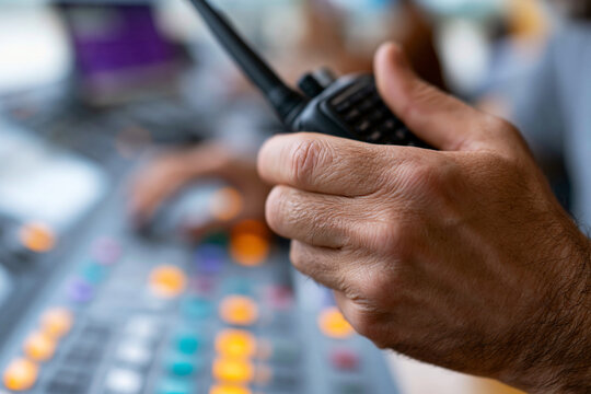 Hand grips radio in focused control room setting. Fingers tense, ready for communication or coordination. Control panel with colorful buttons glows softly behind