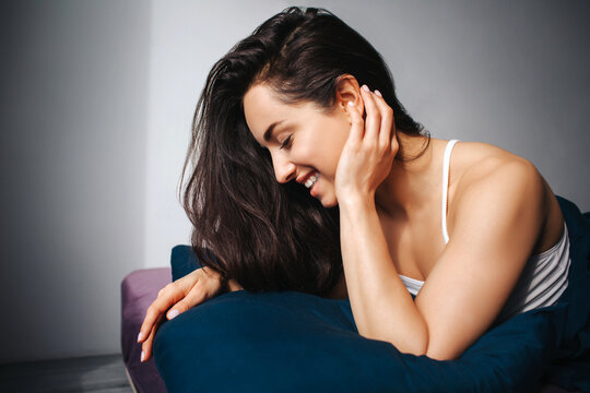Young beautiful woman in morning bed at home. Cheerful hot sexy attractive feminine model on picture smiling. Look down and touch her ear.