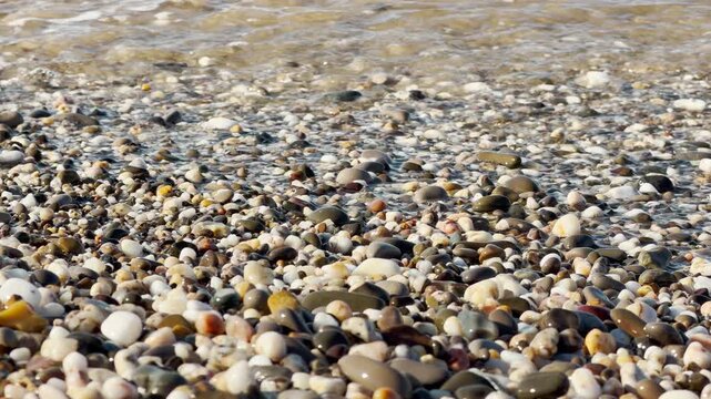 Clear ocean water flows over a bed of polished, colorful shoreline stones. The gentle waves push and pull the pebbles, creating a soft, rhythmic clacking and rushing sound.