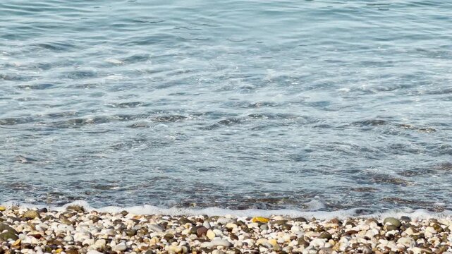 Clear sea water rolls over the shore and turns into white foam. The waves pull back across the small pebbles, creating a soft swishing and fizzing sound.