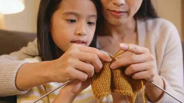 Placing hands, mom in beige cardigan guiding daughter learning knitting on couch with yarn, needles