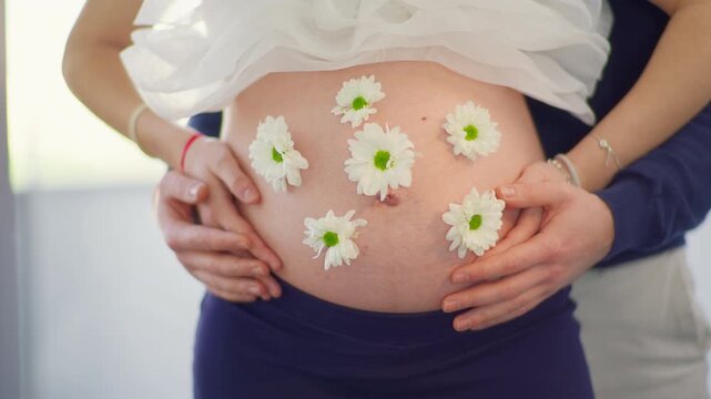 White daisy flowers arranged on pregnant belly while partner places both hands gently from behind, bright indoor light background