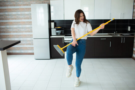 Young woman on kitchen during quarantine. Holding broom in hands as musical instrument and pretend to play on it. Have fun alone after cleaning floor.