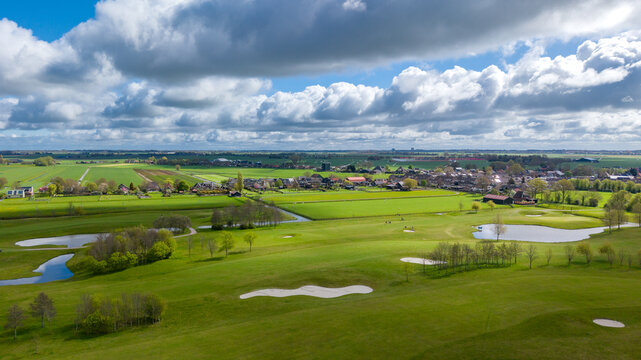 Aerial view of a lush green golf course with sand bunkers and ponds next to a rural village under a dramatic cloudy sky in Netherlands.