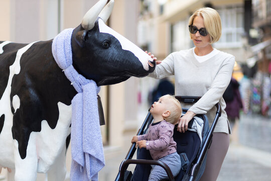 CuriouMother and little son exploring city streets and discovering a funny cow sculptures toddler in a stroller looking at a cow statue in front of a shop in Spain.