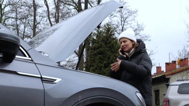 Frustrated woman standing by her broken down car with an open hood, anxiously calling for help on her smartphone while stranded on the side of the road in cold weather during an emergency