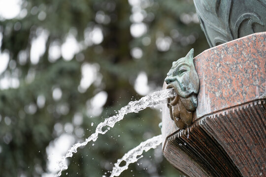  Classical city fountain detail with a decorative bronze mascaron and splashing water.  Bronze gargoyle head water spout on a granite fountain, outdoor park background.