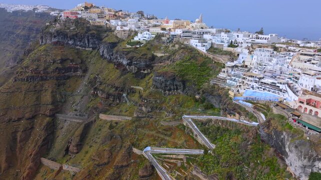 A dramatic high-angle aerial video of the famous zigzagging stone staircase in Fira, Santorini, descending the steep volcanic cliffs from the white village down toward the Aegean Sea.