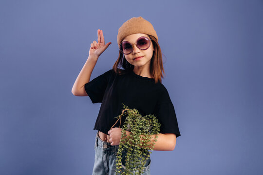 Young girl with brown hair wearing oversized sunglasses and a beige beanie poses playfully while holding a potted plant against a solid purple background