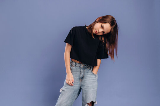 Young woman with long brown hair wearing a black cropped t-shirt and ripped jeans poses against a solid light purple background in a studio setting