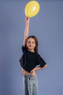 Young girl with long brown hair wearing a black crop top holds a yellow balloon high above her head against a solid blue background