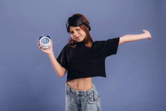 Young girl wearing black crop top and denim jeans holds alarm clock while stretching her arm against a solid blue background in a playful pose