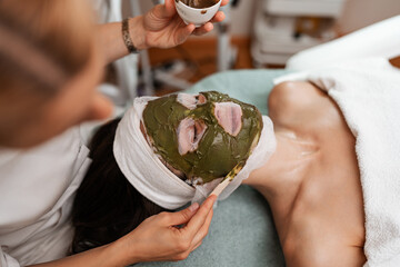 A professional esthetician applies a natural green algae and seaweed facial mask over gauze onto a woman's face during a rejuvenating spa treatment. © Dusko