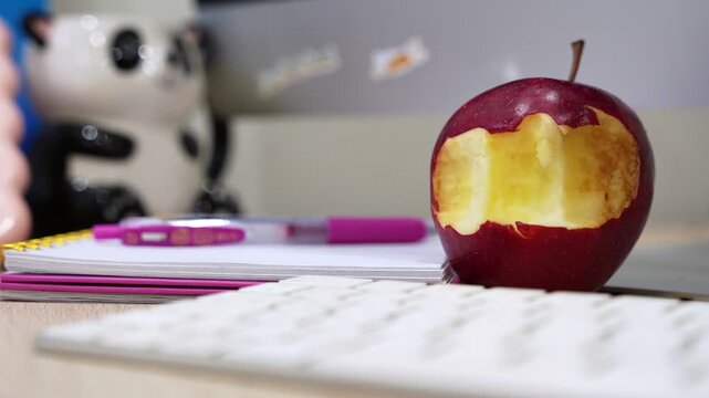 a bitten red apple sitting on a wooden desk next to a notebook and keyboard. A healthy snack choice in a modern office environment with bright indoor lighting.