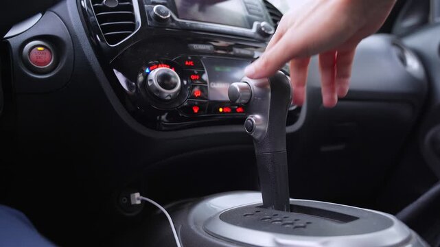 a driver shifting an automatic gear lever inside a modern vehicle cabin. The dashboard with climate controls is visible in the blurred background under natural daylight.