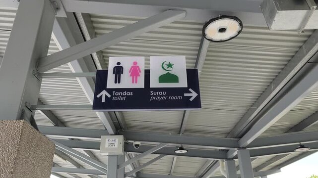Bilingual signage in Malay and English directs users to restrooms and a prayer room. Simple icons ensure clarity. The sign hangs from a high structure for maximum visibility.