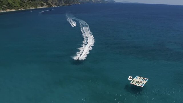 Aerial drone view of speedboat moving across clear blue sea leaving curved wake near rocky coastline with hills and calm water under bright daylight sky