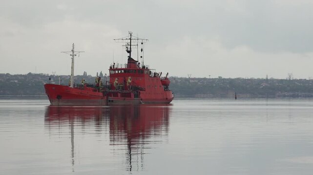 Wide shot of bright orange work ship with deck cranes on still mirror water of a port estuary under cloudy gray sky with distant shoreline in background