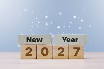 Wooden cubes displaying the phrase New Year 2027 on a wooden table against a blue background with sparkling snow. Concept annual planning, holiday greetings and upcoming year goals. © vadish