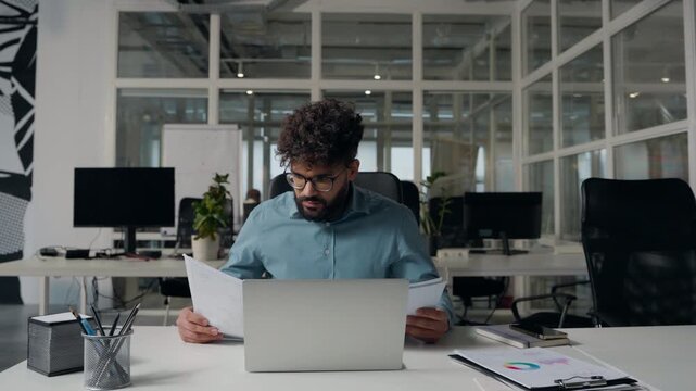 Busy stressed Arabian business man analyzing papers compare data in laptop paperwork difficulty deadline document review at office table ethnic businessman working online male managing finance analyze