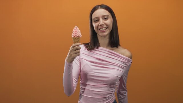 Woman holding ice cream cone and smiling in studio with pink soft serve, casual top and visible hand gripping waffle cone; joy playfulness.