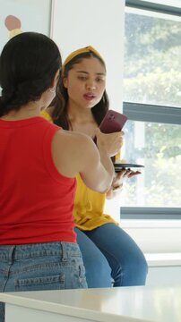 Vertical video: Standing red-top showing maroon phone to yellow friend at kitchen counter laughing