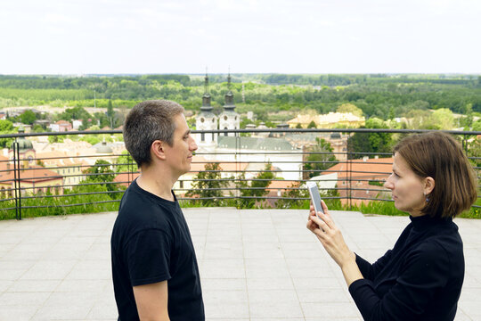 Woman photographing male partner with smartphone on scenic overlook above historic city. Portrays candid vacation documentation, personalized trip keepsakes, and experience-centered travel narration.