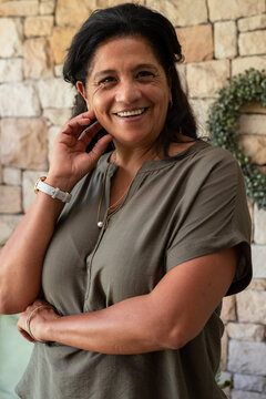 Mature woman smiling while touching ear and wearing olive-green blouse on porch with pearl pendant