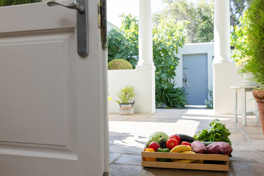 Wooden crate of produce is sitting on tiled porch near open white door, sunlight casting shadows