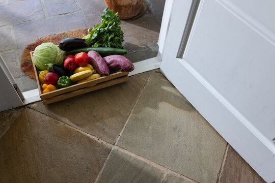 Wooden crate is sitting on stone tiles near open white door, holding cabbage, eggplant, cucumbers