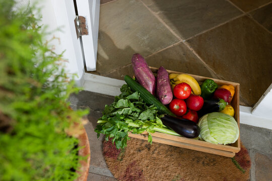 Wood crate sitting on coir mat at slightly open door, overflowing with cabbage, eggplant, cucumber